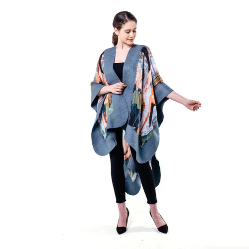 A woman modeling a blue-gray open-front poncho with a multicolor abstract print, worn over a black top and black leggings against a plain white background.