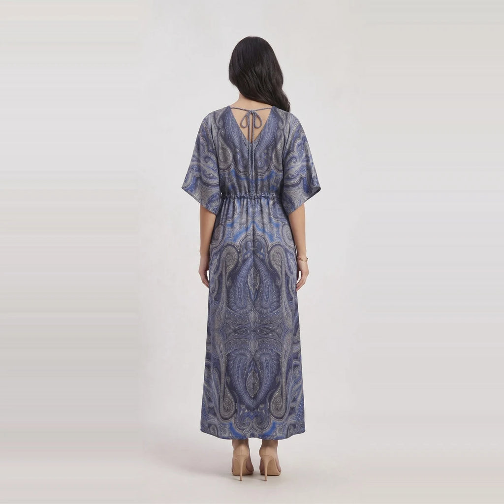 A woman viewed from the back wearing a loose, floor-length dress with short wide sleeves and a paisley-style print in blue, gray, and white. The dress has a tie detail at the back neckline and a gathered waist.