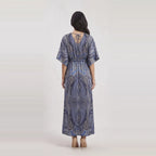 A woman viewed from the back wearing a loose, floor-length dress with short wide sleeves and a paisley-style print in blue, gray, and white. The dress has a tie detail at the back neckline and a gathered waist.