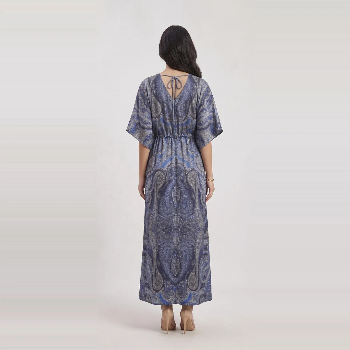 A woman viewed from the back wearing a loose, floor-length dress with short wide sleeves and a paisley-style print in blue, gray, and white. The dress has a tie detail at the back neckline and a gathered waist.
