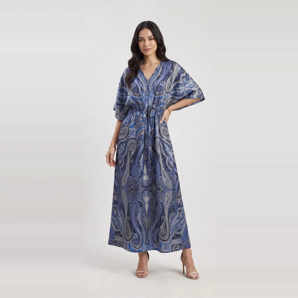 A woman modeling a blue and gray paisley-patterned maxi dress with a V-neck, short wide sleeves, and a drawstring waist, set against a light gray studio background.