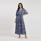 A woman modeling a blue and gray paisley-patterned maxi dress with a V-neck, short wide sleeves, and a drawstring waist, set against a light gray studio background.