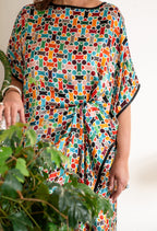 A person wearing a loose, short-sleeve  silk dress with a colorful geometric mosaic pattern, featuring black trim along the neckline and sleeve edges. 