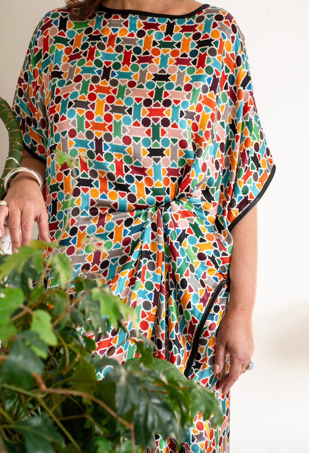 A person wearing a loose, short-sleeve  silk dress with a colorful geometric mosaic pattern, featuring black trim along the neckline and sleeve edges. 